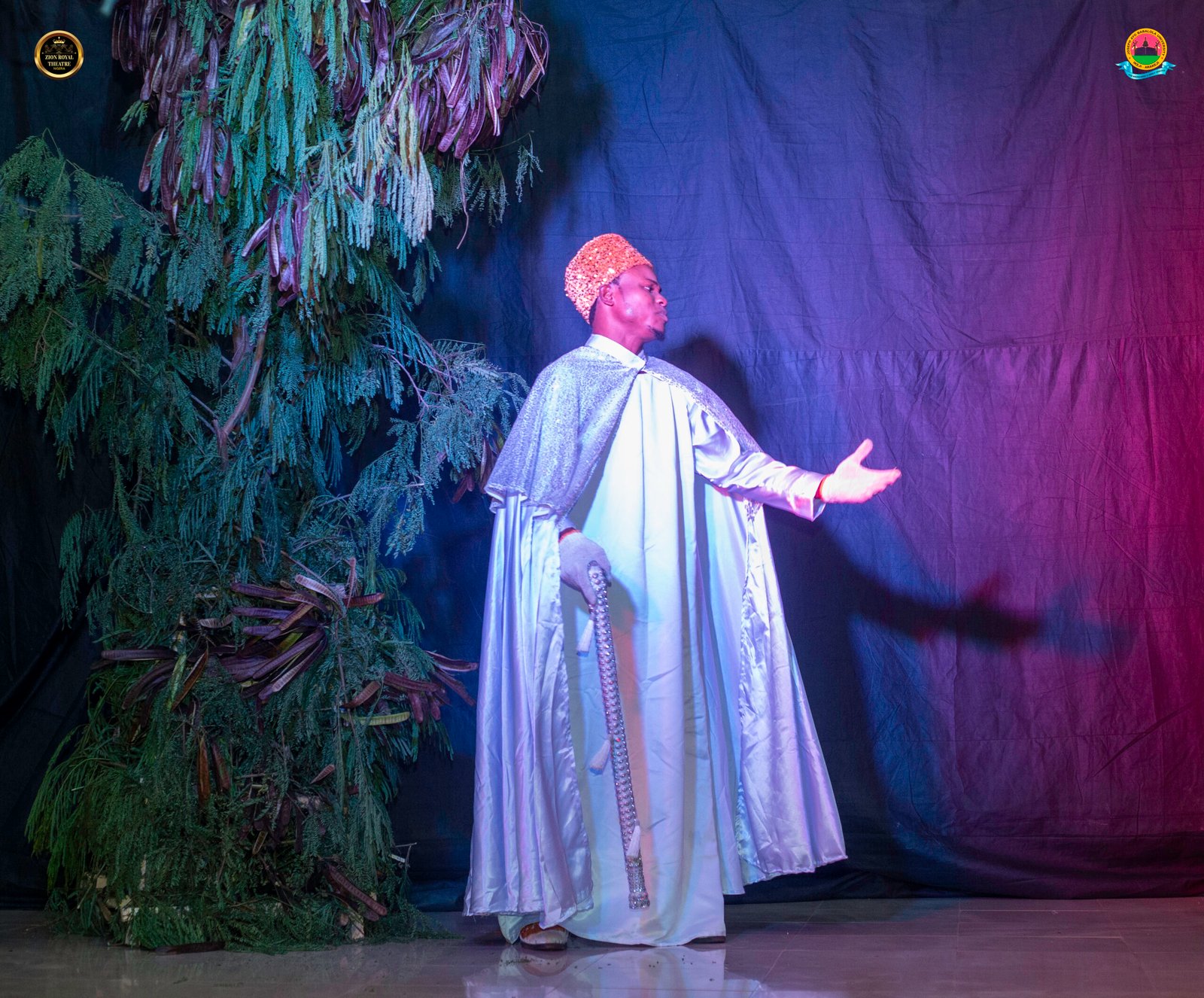 A man wearing a white gown with on stage with bright and red light behind the man - Zion Royal Theatre